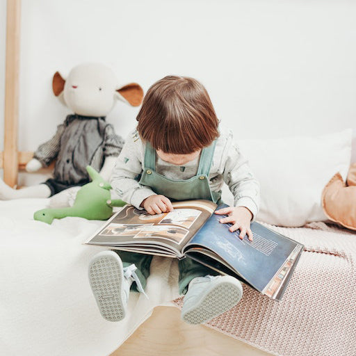 Young girl reading a picture book on her bed surrounded by soft toys. 